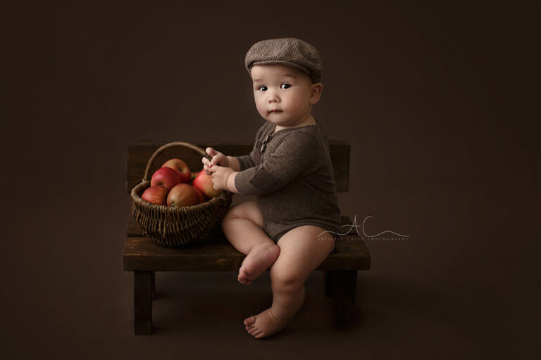 Bromley Baby Portraits | Louie 3 Bromley Baby Portraits | 7 months old baby boy sits on a small wooden bench and holds a wicker basket full of red apples