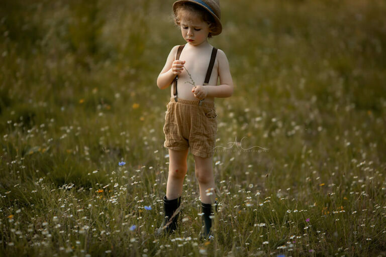Beautiful London Child Photography in Wildlower Field | portrait of a 4 year old boy wearing a straw hat and looking at the small wildflower
