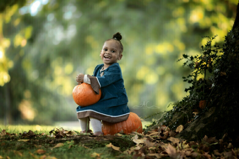 Best Autumn London Toddler Photo Session | 2 year old girl sits on a massive pumpkin and holds a smaller pumkin in her hands