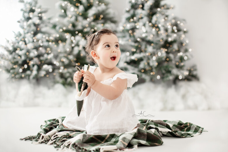 London 2023 Magical Christmas Mini Sessions | excited 1 year old baby girl playing with Xmas toys with snowy Xmas trees in the background