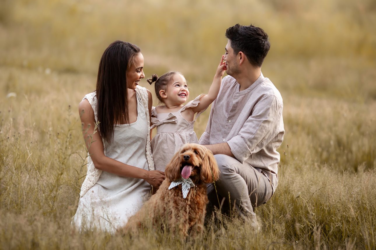 London Summer Mini Sessions | 2 year old girl playing with her parents and a dog in a long grass field during a summer photo session