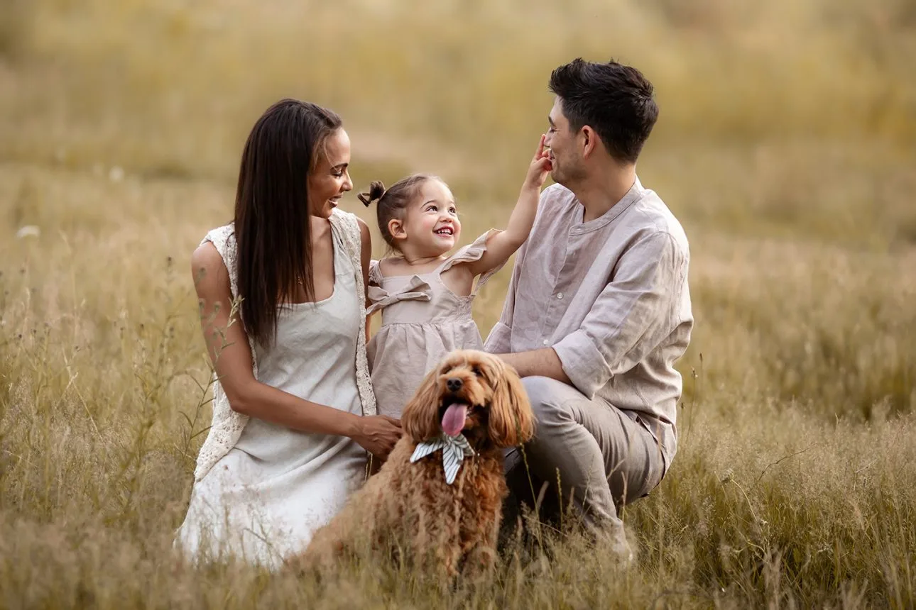 London Summer Mini Sessions | 2 year old girl playing with her parents and a dog in a long grass field during a summer photo session