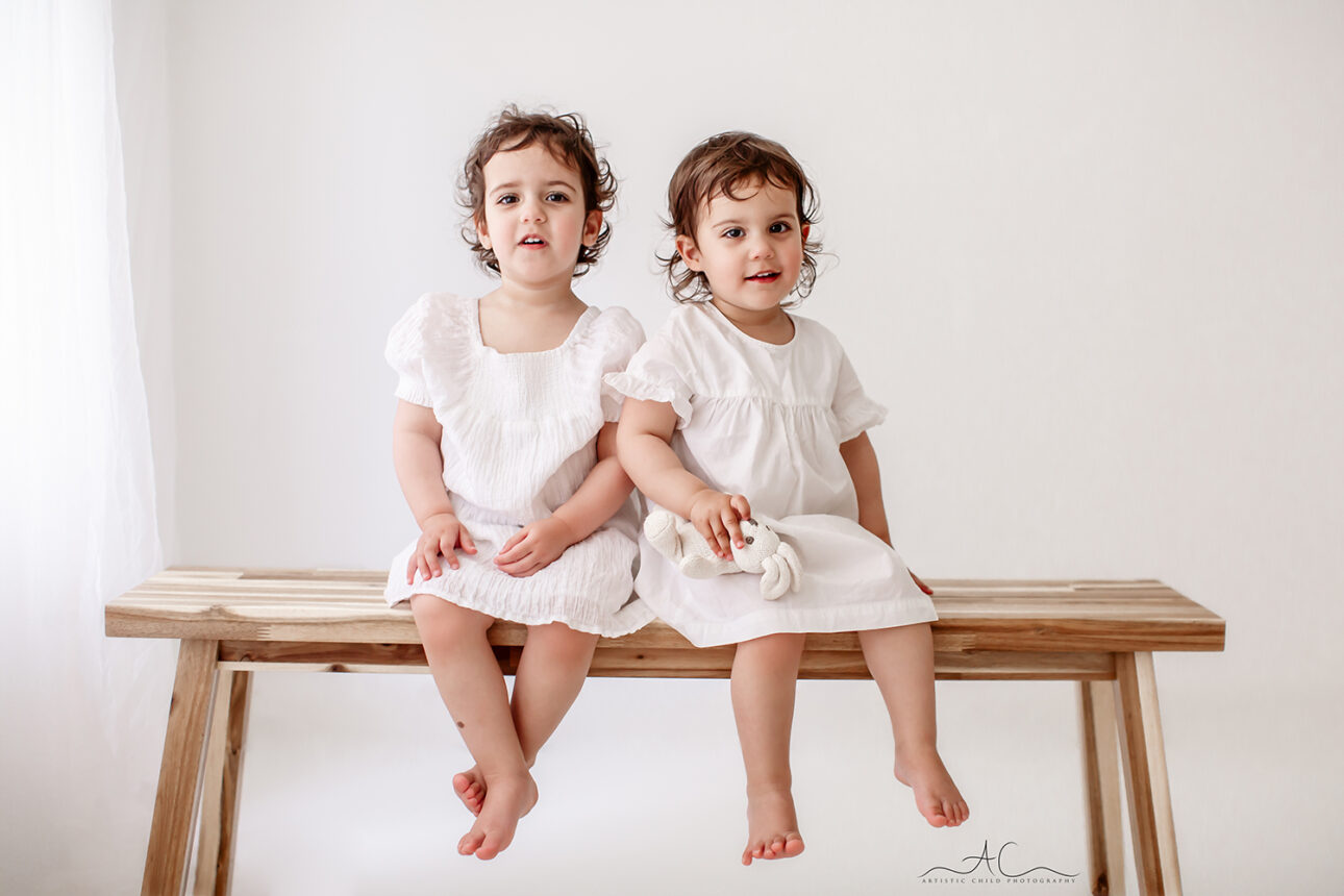 London Twin Toddler Girls Photoshoot | portrait of 2 year old twin sisters sitting on a bench and smiling