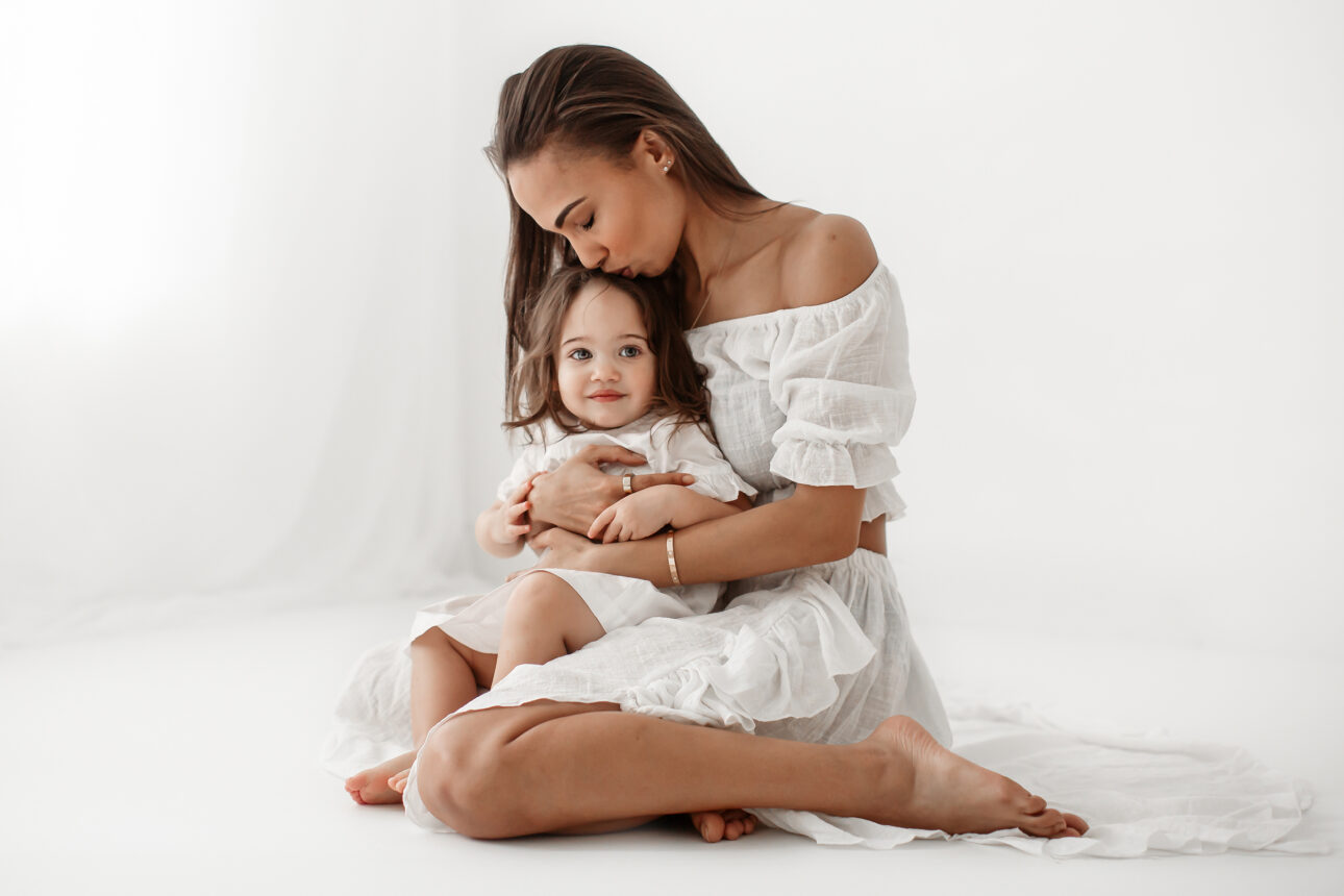 London Mummy and Me Photography | Intimate portrait of mother kissing her toddler daughter while holding her on her lap during London mummy and me photo session in South East London studio